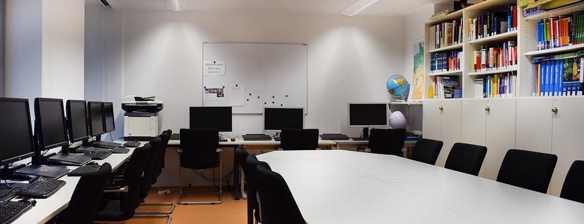 Sie sehen eine Aufnahme des Geomedien-Lernlabors. Mittig im Raum befindet sich ein großer Konferenztisch, seitlich stehen PC-Arbeitsplätze. 
